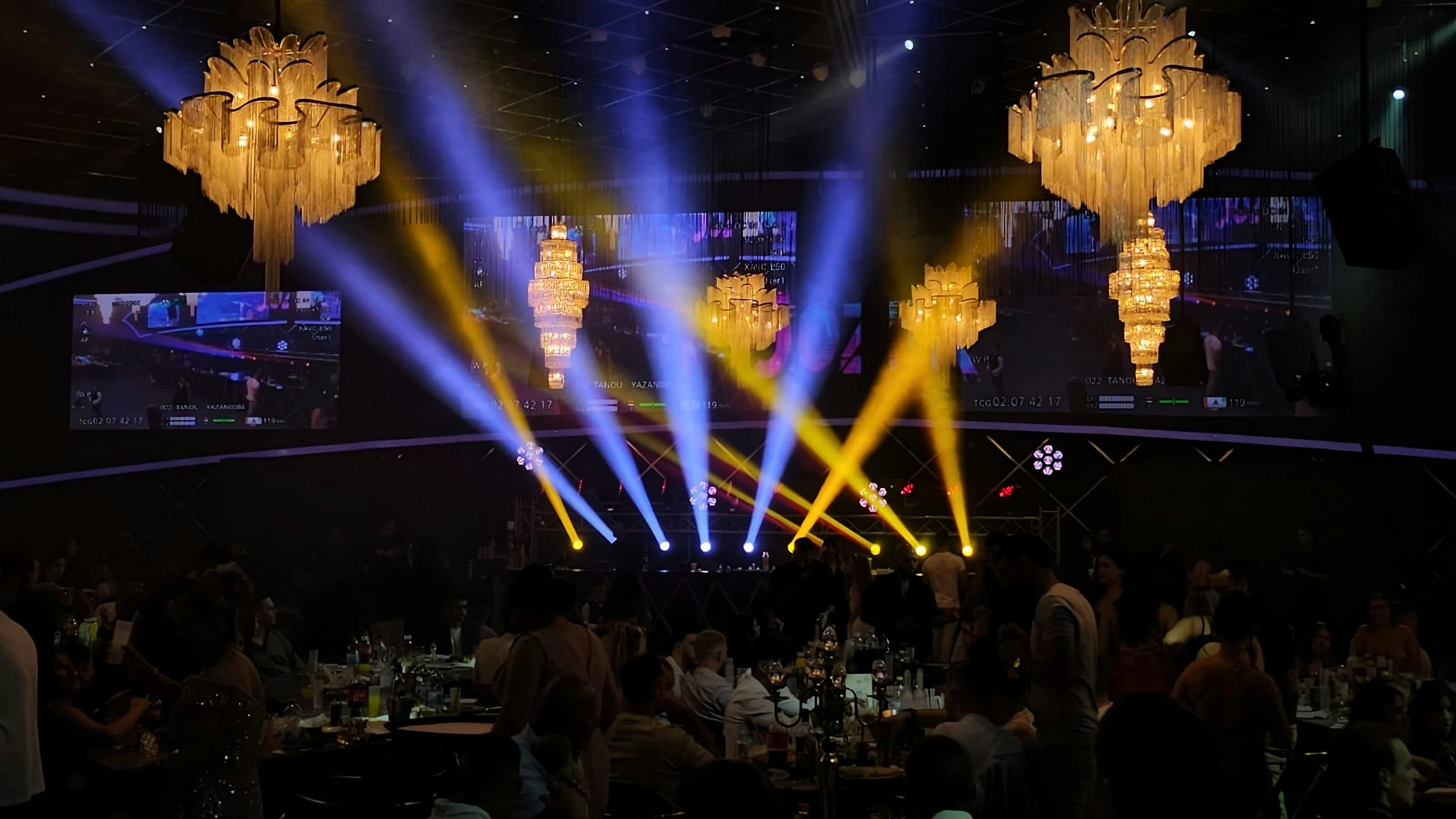 Guests sit at tables under golden chandeliers with blue and yellow light beams on Awards Night stage