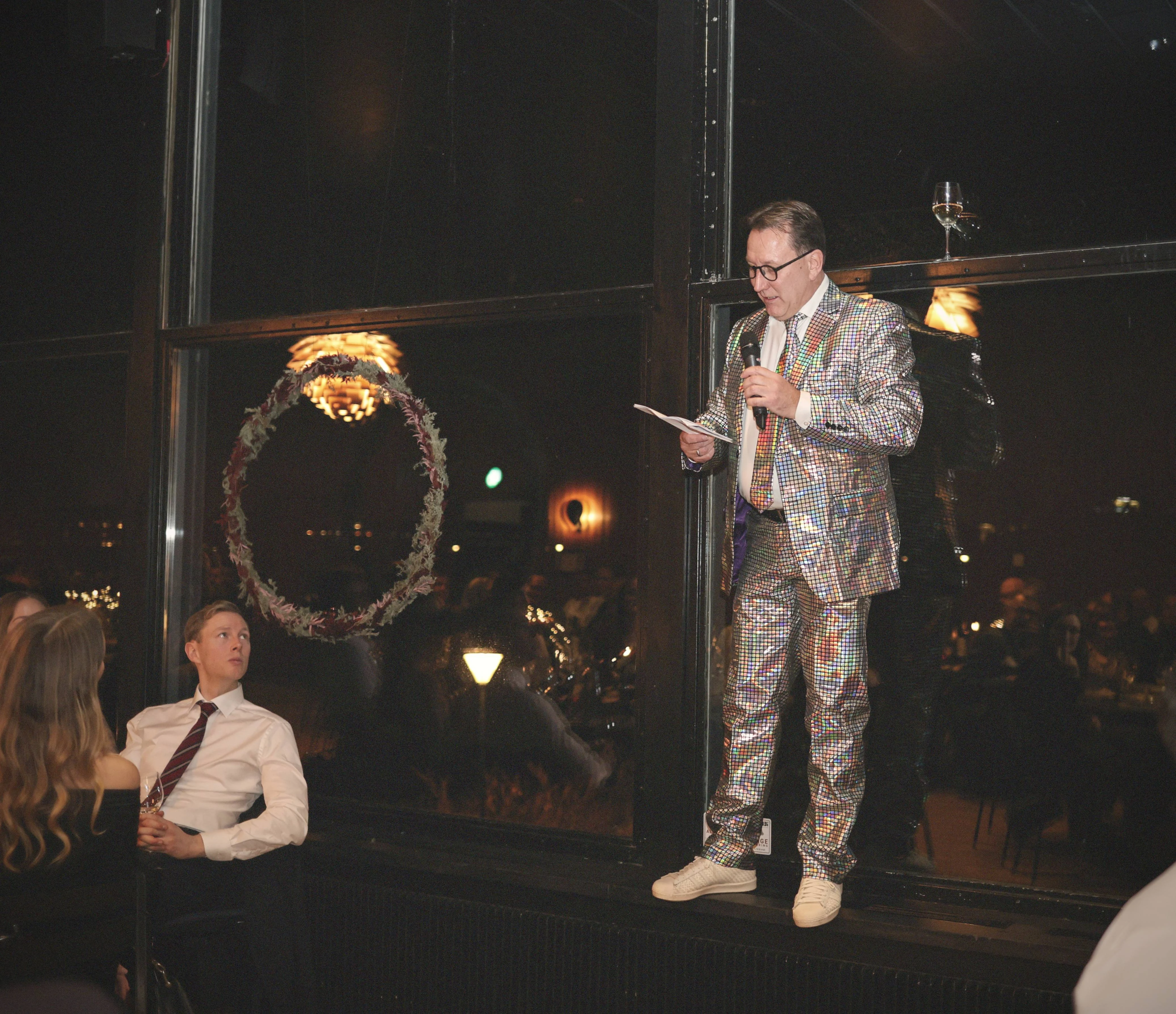 Persoon in glitterend pak spreekt met microfoon op verhoogde plek tijdens feestelijk evenement