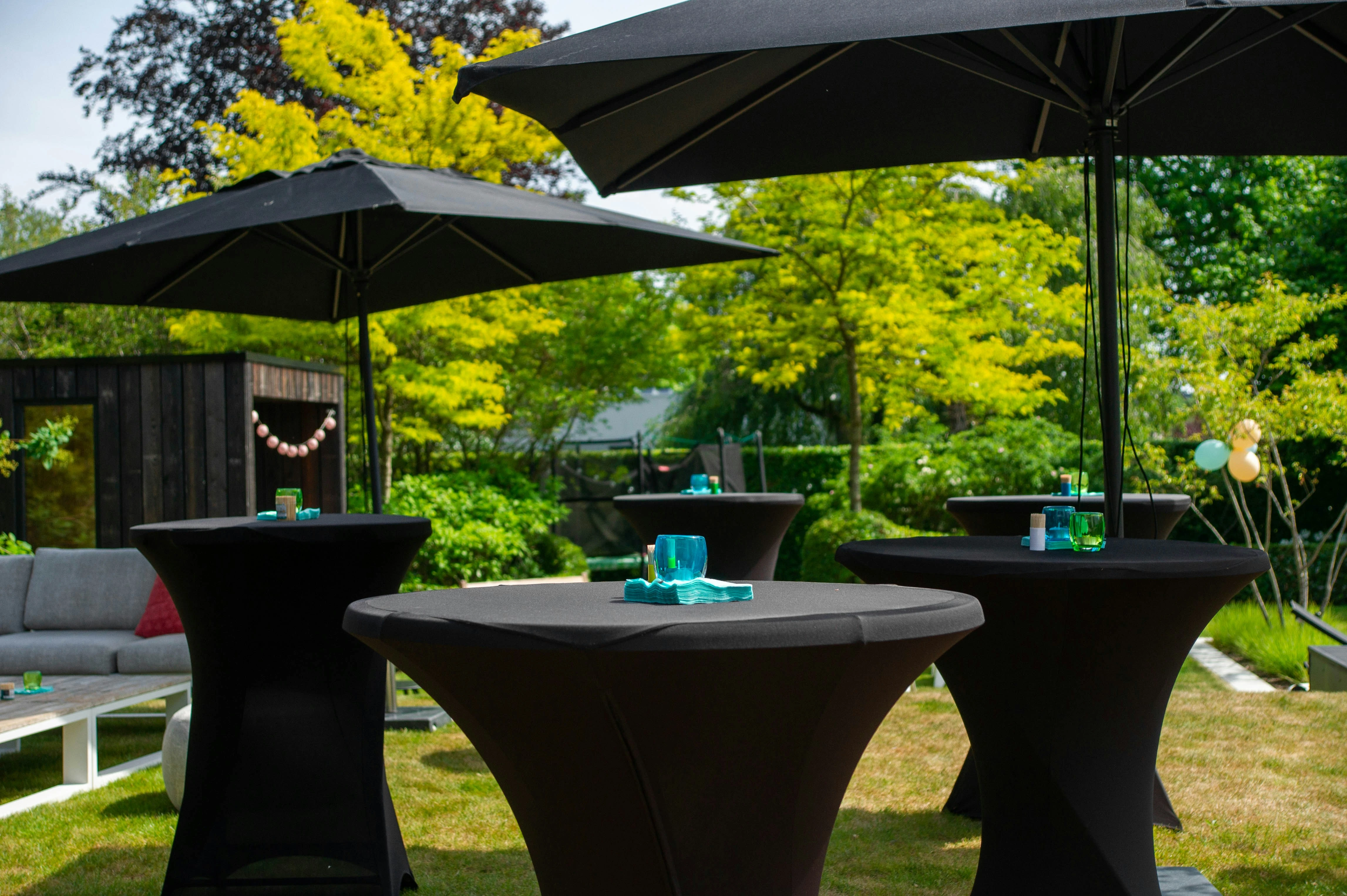 Tuinfeest op gazon onder zwarte parasols met staande tafels en groene bebossing